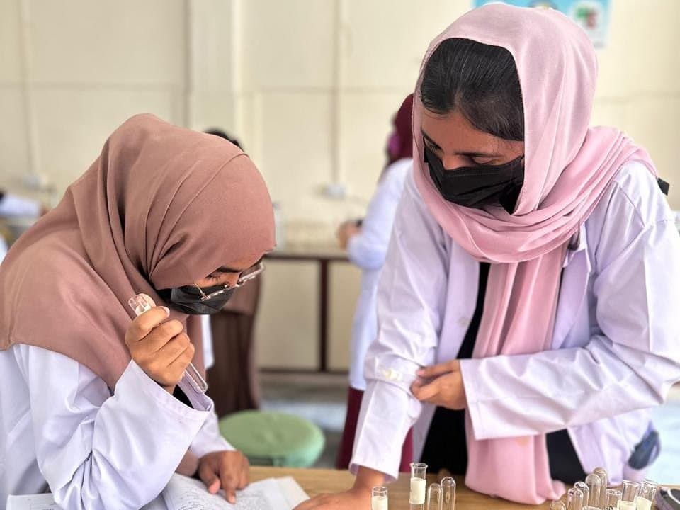 Two ISSA students in white coats recording results beside test-tube racks during a laboratory practical