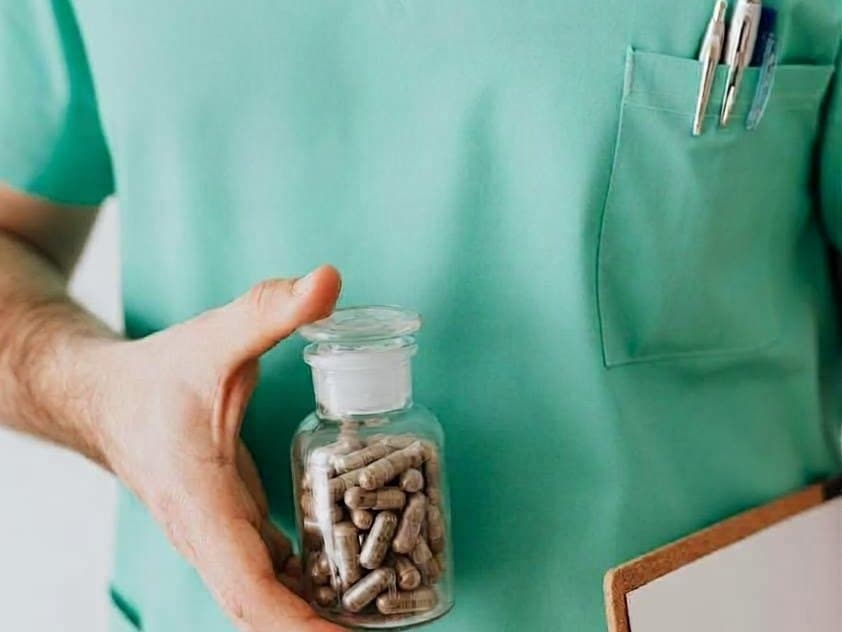 A pharmacy technician in green scrubs holding a glass jar of capsules, with pens in the pocket and a clipboard