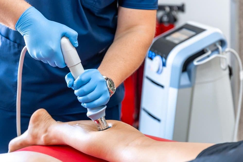 A physiotherapist in blue gloves treating a patient's calf with a shockwave therapy device