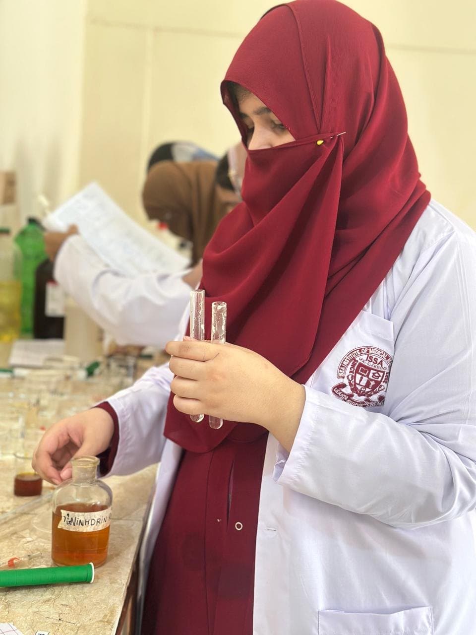 An ISSA pharmacy student in a white coat holding test tubes at the laboratory bench during a practical